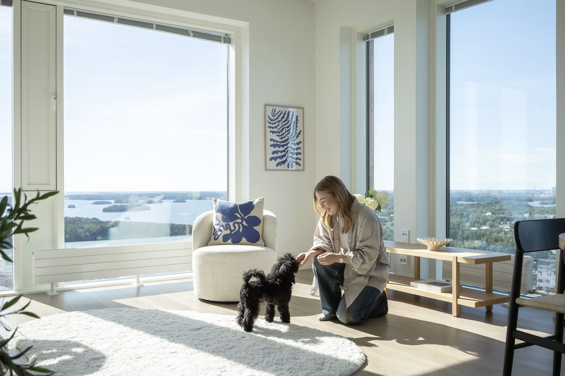 A woman playing with a small black dog in a bright, modern apartment with large windows and scenic views.